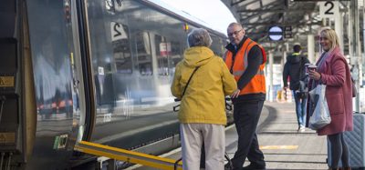 Staff member assisting passenger with mobility aid onto train with ramp deployed at a train station